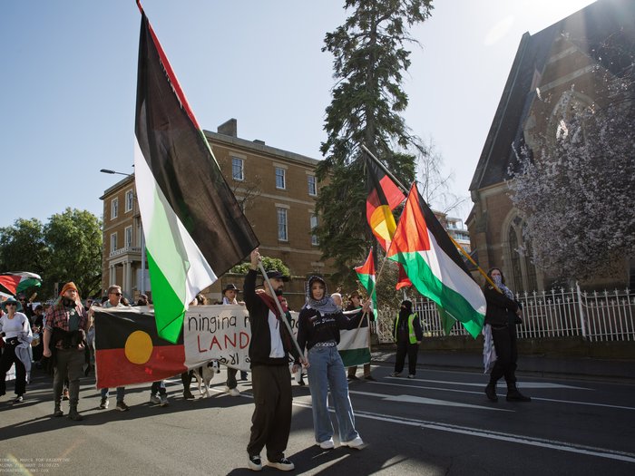 A man and a woman holding two big Palestinian flags. Next to them someone is holding a big Aboriginal flag. Behind them a large group of people is marching. A banner in the front reads 'land back' in Palawa kani, Arabic and English.