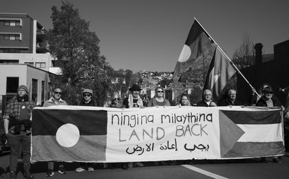 A black and white photo of people holding a banner that features both the Aboriginal flag and the Palestinian flag and says 'land back' in Palawa kani, Arabic and English. Someone is also holding a flag pole with both an Aboriginal and Palestinian flag.