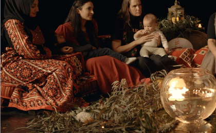 three women sitting together. One is holding a baby and a toddler appears to be sleeping in the arms of another one of them. Eucalyptus leaves can be seen on the ground.