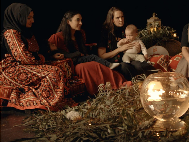 three women sitting together. One is holding a baby and a toddler appears to be sleeping in the arms of another one of them. Eucalyptus leaves can be seen on the ground.